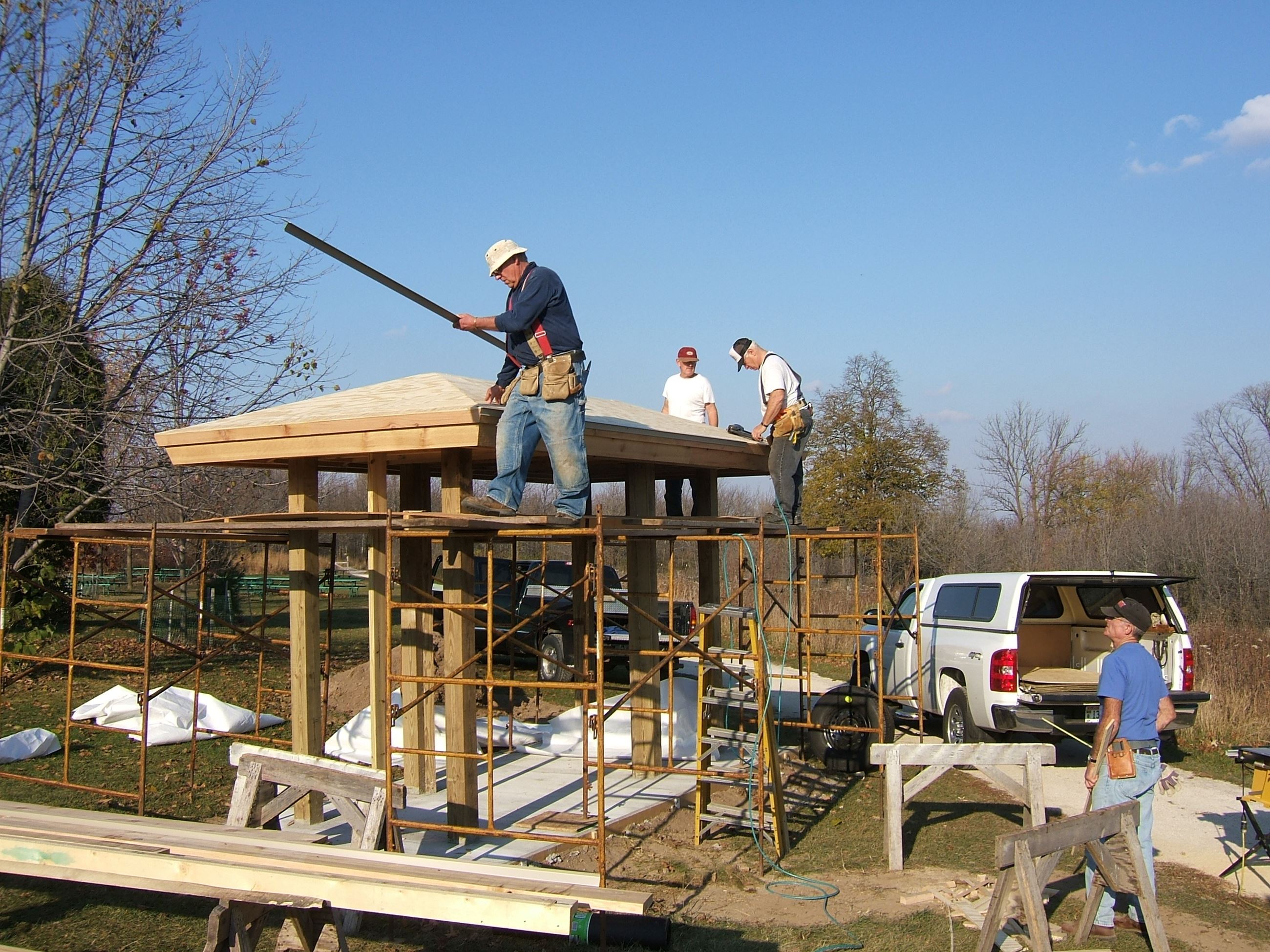 Four me building a kiosk. 3 are standing on scaffolding and working on the roof, 1 is on the ground 